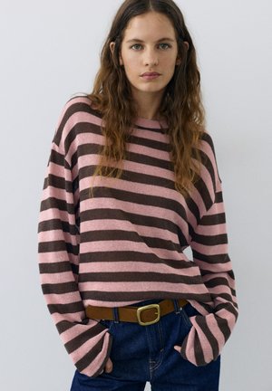 Jeune femme aux longs cheveux ondulés portant un pull ample rayé rose et marron, un jean bleu et une ceinture marron, debout les mains dans les poches.