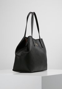 Sac en cuir noir texturé avec double sangle, base structurée et petit logo doré à l'avant, expos é sur une surface blanche.