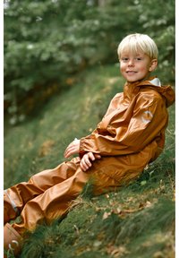 Hnědý nepromokavý pláštěnkový komplet s elastickými manžetami a kapucí, s bílým detailem na rukávu. Dítě sedící na travnatém terénu.