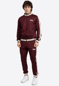Maroon trenerka od glatkog materijala; sadrži bijele detalje, jaknu na patentni zatvarač, logo na prsima i hlačama te rebraste manšete.