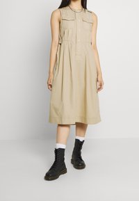 Beige, hihaton mekko, jossa edessä napitus, kaksi rintataskua, levenevä helma ja väljä istuvuus. Yhdistetty mustiin nauhallisiin saappaisiin.