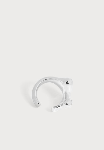 Une bague manchette ouverte en argent poli, ornée d’une petite pierre triangulaire noire, sur un fond clair et uni.