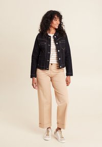 Donkerdenimjack met gouden stiksel, twee borstzakken, gecombineerd met een crèmekleurig en beige gestreept shirt en beigekleurige wijde broek. Witte sneakers.