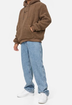 Haut en polaire marron avec une poche avant, capuche avec cordon de serrage, et poignets côtelés, associé à un jean décontracté bleu clair et des baskets blanches.