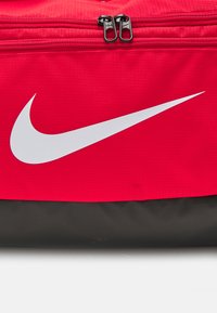 Rød og sort Nike træningstaske lavet af tekstureret stof, der har et fremtrædende hvidt swoosh-logo og to sorte lynlåsgreb.