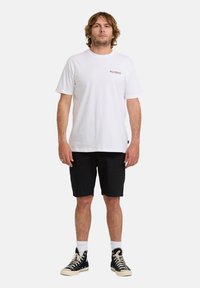 Ein Mann steht mit dem Gesicht nach vorne, trägt ein weißes Billabong-T-Shirt, schwarze Shorts, weiße Socken und schwarze High-Top-Sneaker.