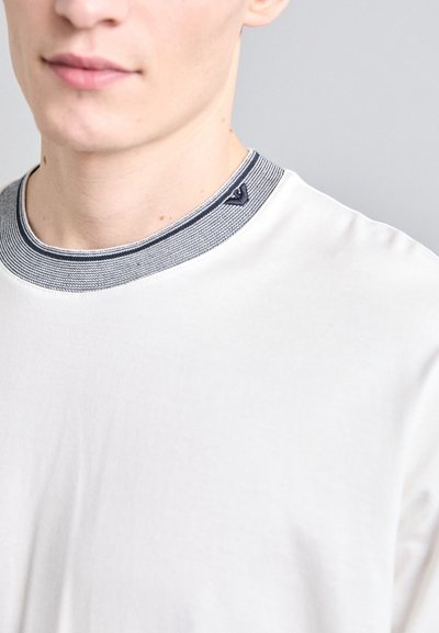 Balta marškinėliai su tamsiai mėlynu ir pilku banguotu apykaklė, kuriuose yra mažas logotipo akcentas kairėje kaklo pusėje. Minkšta tekstūra.