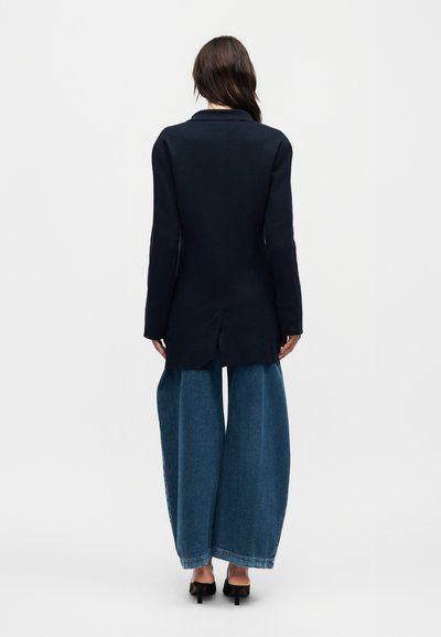 Γυναίκα με την πλάτη να κοιτάζει, φοράει ένα μακρύ μαύρο σακάκι, φαρδιά μπλε τζιν και μαύρα ψηλοτάκουνα παπούτσια σε ένα απλό φωτεινό φόντο.