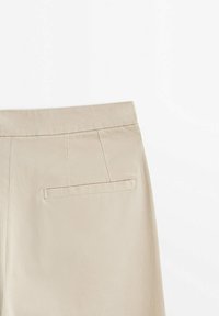 Pantaloni beige vista posteriore che mostrano la vita, i dettagli delle cuciture e una tasca a filetto singola sul lato destro, il tutto su uno sfondo bianco.