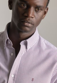 Camisa de algodón abotonada con estrechas franjas burdeos y blancas, cuello con puntas y un pequeño logo en el pecho. Textura suave.