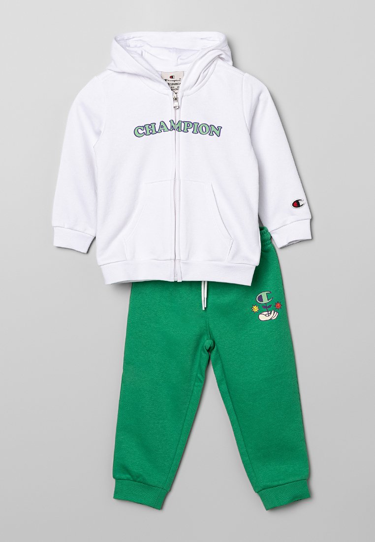 Felpa bianca con zip e scritta "CHAMPION" in verde e viola, abbinata a pantaloni da tuta verdi con logo Champion e grafica di una mano colorata.