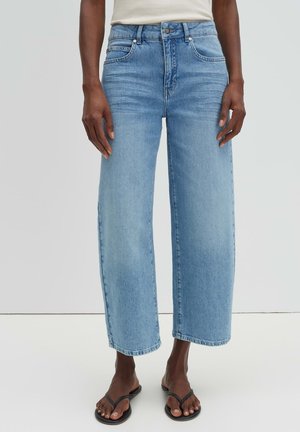 Personne portant un jean large bleu clair coupé au-dessus de la cheville et des sandales noires à lanières, debout devant un fond uni clair.