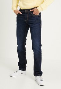 Donkerblauwe jeans met contrasterende oranje stiksels, slim fit, vijf-pocketdesign en riemlussen, gedragen met witte sneakers en een gele hoodie.