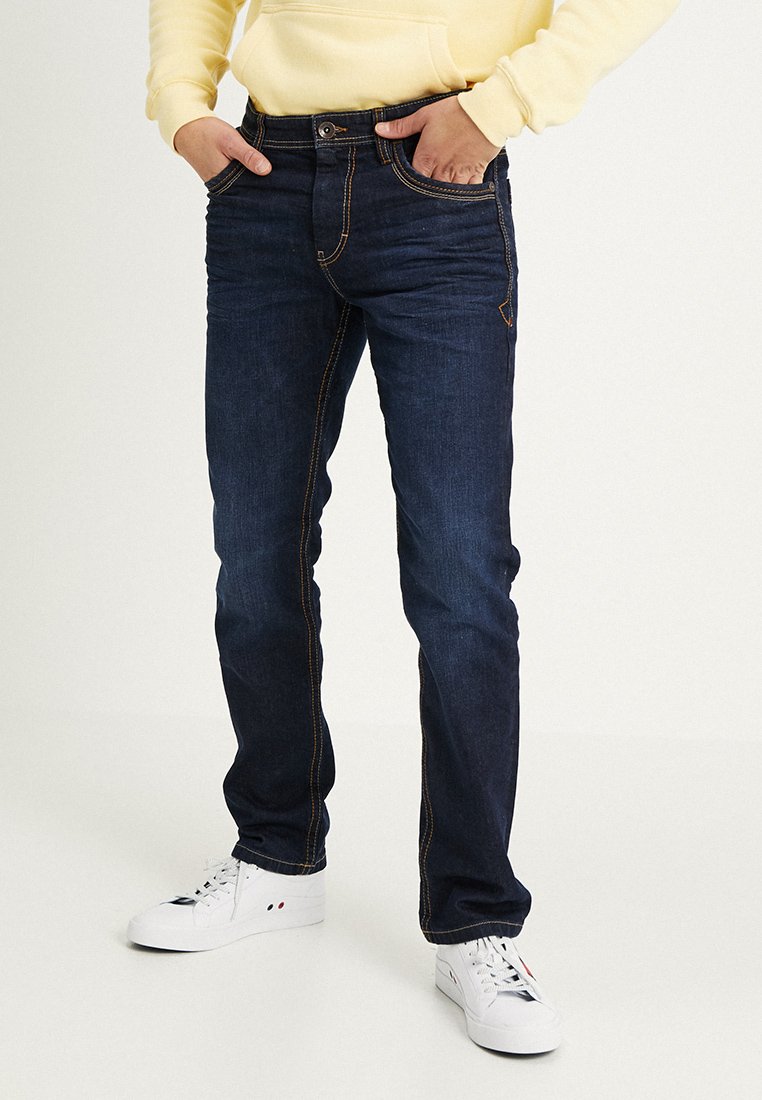 Donkerblauwe jeans met contrasterende oranje stiksels, slim fit, vijf-pocketdesign en riemlussen, gedragen met witte sneakers en een gele hoodie.