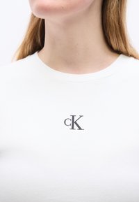 Γυναίκα που φοράει λευκό μπλουζάκι με στρογγυλή λαιμόκοψη και μικρό μαύρο λογότυπο "cK" κεντραρισμένο κάτω από το πηγούνι της, με φόντο ένα απλό λευκό τοίχο.