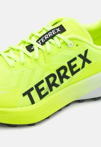 Neon geltonos spalvos sportinė avalynė su tinkline viršutine dalimi, juodu "TERREX" prekės ženklu ir kontrastinga pilka išorine sola. Yra plokščios raišteliai ir sustiprintas pirštas.
