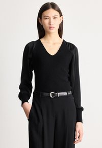 Sieviete ar taisniem tumšiem matiem, valkājot melnu V veida kakla džemperi ar uzpampušām piedurknēm, melnu jostu un melnas bikses, stāv ar vienu roku kabatā.