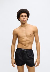 Fekete boxeralsó logós derékpánttal, gombos záródással és sima, fényes textúrával. A modell póló nélkül áll.