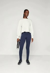 Haut court à manches longues gris clair associé à des leggings navy. Le mannequin tient une bouteille d'eau noire et porte des chaussures de sport noires. Textures lisses.