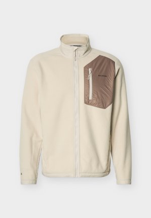 Bézs teljes hosszúságú cipzáros polárdzseki magas gallérral, bal oldalon barna cipzáras mellzsebbel, melyen Salomon logó látható.