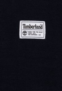 Tissu tricoté marine avec une étiquette blanche arborant le logo "Timberland", un graphique d'arbre et le texte "CONSTRUIT POUR LES AUDACIEUX" avec des informations de localisation.