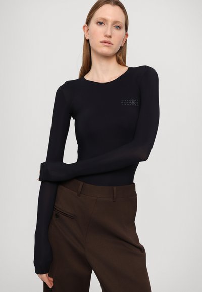 Black long-sleeve bodysuit with a fitted design, featuring a subtle graphic on the chest, paired with brown tailored trousers.