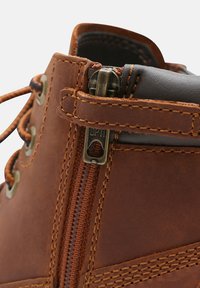 Stivale alla caviglia in pelle marrone con chiusura a cerniera e lacci, caratterizzato da accessori in ottone, dettagli di cucitura e texture liscia.