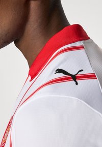 Camisola de desporto branca com detalhes e gola vermelhos. Apresenta um logótipo preto da Puma e riscas vermelhas horizontais no ombro. Tecido texturizado.