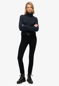 Jersey de cuello alto de punto trenzado en color marino, jeans ajustados negros y botas de combate negras con cordones. El patrón texturizado en el jersey añade interés visual.