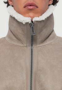 Beige színű, shearling galléros dzseki, teljes zipzárral, textúrált velúr anyagból, sima felülettel. Figyelemre méltó részletei közé tartozik a fém zipzár szerelvény.