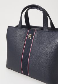 Tummansininen tekokoiran nahkakassi, jossa on kaksi lyhyttä käsinettä, keskeinen punavalkoinen raita ja kultainen emblem etupuolella.