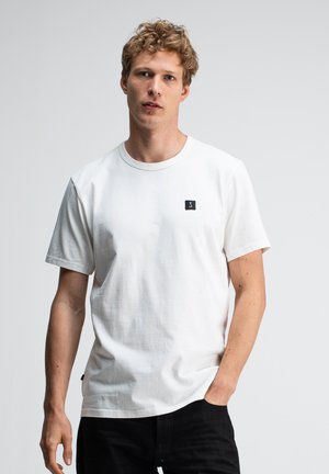 Junger Mann mit lockigem Haar, der ein schlichtes weißes T-Shirt mit kleinem schwarzem Logo trägt, entspannte Hände, neutraler Gesichtsausdruck, schlichter hellgrauer Hintergrund.