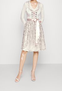 Robe à corsage lacé avec de longues manches texturées, une ceinture en satin rose, et une broderie complexe sur une jupe légère et fluide.