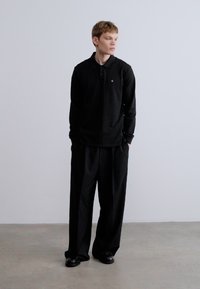 Mustad pika varrukaga polosärk kraega, millel on väike logo. Lahtise istuvusega mustad püksid, kombineeritud mustade kingadega, neutraalsel taustal.