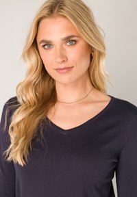 Marineblauwe V-halstrui met lange mouwen, vervaardigd uit een gladde, textuurrijke stof. Accenten zijn onder andere een delicate gouden ketting en zacht, golvend blond haar.