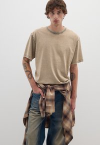 T-shirt in cotone beige con collo a giro, dettagli di cucitura sottili, abbinato a jeans azzurri chiari e a una camicia in flanella a quadri marrone legata in vita.
