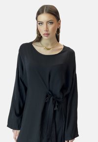 Zwarte lange jurk met lange mouwen, gemaakt van textuurstof, met een ronde hals en een stoffen strik in de taille. Versterkt met gouden sieraden.