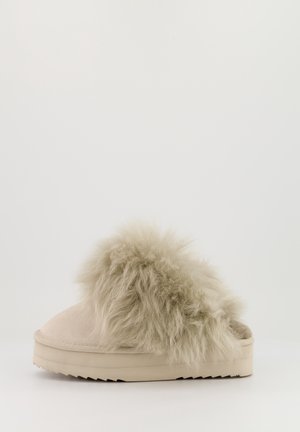 Chausson beige à enfiler avec semelle épaisse et fourrure douce et pelucheuse recouvrant la partie supérieure, présenté sur un fond clair uni.