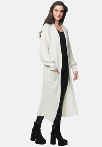 Cardigan lungo in avorio realizzato in tessuto a maglia testurizzato. Presenta un frontale aperto, due tasche anteriori e dettagli a costine sulle maniche. Abbinato a un top e pantaloni neri.