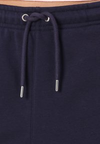 Tmavě modré tepláky mají stahovací pas s kovovými koncovkami a boční švy. Materiál vypadá měkký a má hladkou texturu.