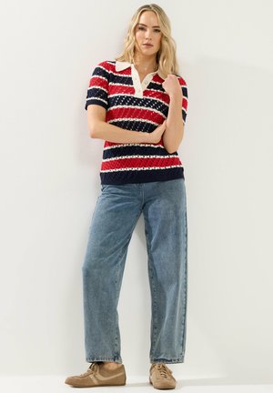 Mujer rubia de pie con una camisa tipo polo de punto a rayas rojas, blancas y azul marino, jeans azules de pierna ancha y zapatillas beige frente a un fondo liso.