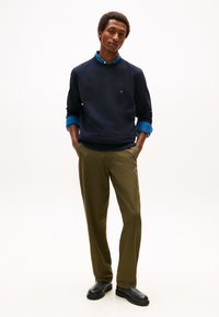 Tamnoplava majica dugih rukava preko plave košulje s ovratnikom, u kombinaciji s maslinasto zelenim hlačama i crnim cipelama; s malim logotipom na prsima.