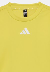 Camisa atlética amarilla hecha de un tejido suave. Diseño de cuello redondo con un logo blanco de Adidas en el pecho. Pequeños acentos blancos en el cuello.