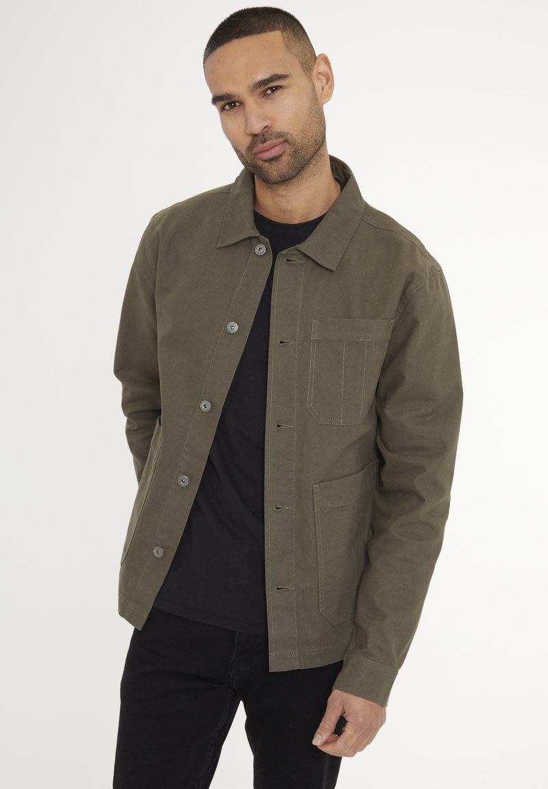 Oliven grøn jakke lavet af bomuld, med en knaplukning, en brystlomme og lige pasform. Båret over en sort skjorte og mørke bukser.