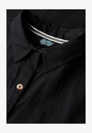 Czarna koszula z kołnierzykiem, wykonana z lekkiego materiału, z zapięciem na guziki po lewej stronie, beżowym guzikiem oraz kontrastową metką z plecionym wzorem wewnątrz kołnierzyka.