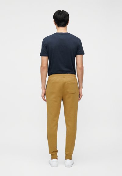 Άνδρας με μουσταρδί παντελόνι jogging, μπλουζάκι σε ναυτικό μπλε χρώμα και λευκά αθλητικά παπούτσια, στέκεται με την πλάτη προς τα εμπρός σε απλό φωτεινό φόντο.