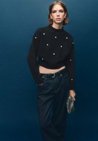 Maglione corto nero con decorazioni in argento, abbinato a jeans in denim scuro a vita alta. Tiene una clutch metallica. Sfondo blu.