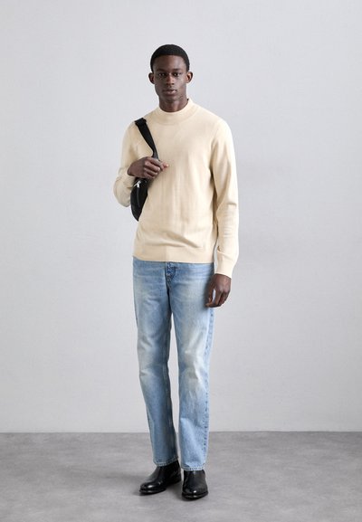 Junger Mann in beigem Pullover, hellblauen Jeans, schwarzen Stiefeln und schwarzer Umhängetasche, der vor einer schlichten, hellgrauen Wand steht.