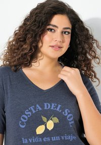 Niebieska koszulka z krótkim rękawem i dekoltem w serek z grafiką cytryny oraz napisem "COSTA DEL SOL la vida es un viaje" w kontrastujących kolorach.