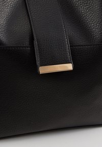 Czarna torebka ze skóry o fakturze, z wyraźnym złotym akcentem z metalu, na którym znajduje się prostokątna plakietka z napisem "ANNA FIELD".